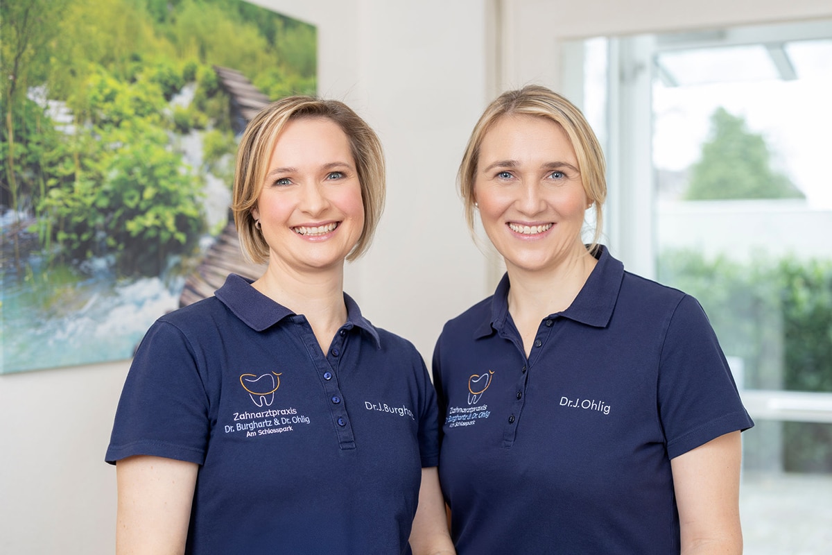 Die Zahnärztinnen Dr. Jeannette Burghartz und Dr. Julia Ohlig präsentieren gemeinsam in einem Portrait-Foto ihre Zahnarzt-Praxis am Schlosspark in Düsseldorf Benrath.